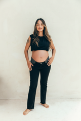 Mujer embarazada de pie usando jeans de maternidad negros con elásticos laterales y ajuste cómodo para el embarazo.